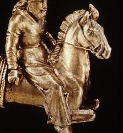 gold carving of a man riding a horse wearing pants and a short tunic with long sleeves