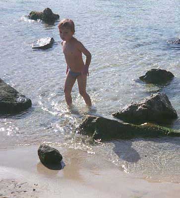 a little white boy wading in the surf on a sunny beach with some rocks