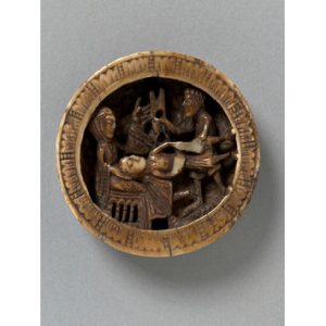 A round piece of brownish ivroy with a woman sitting and a man lying down with his head in her lap. Another man stands in the background - the attendant who brings Delilah the scissors